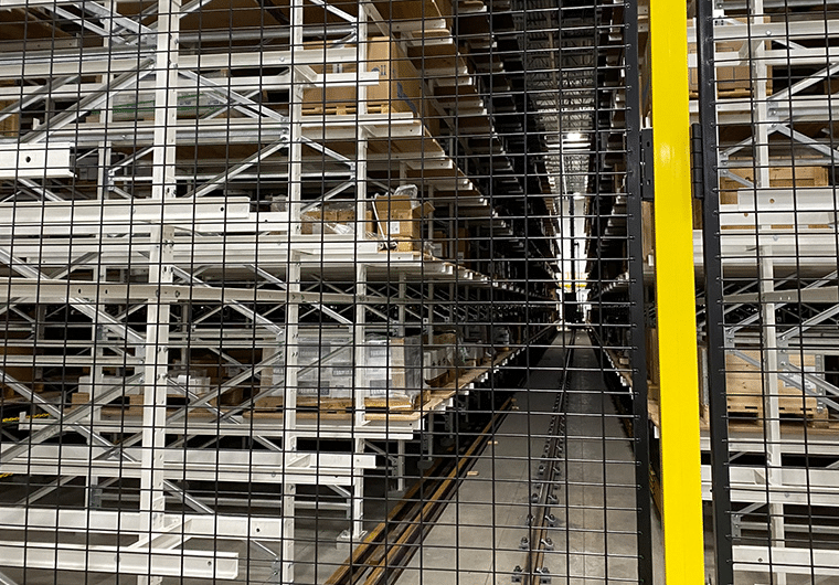 Warehouse aisle with tall metal shelving, pallets stacked on racks, viewed through a metal mesh gate with a yellow post on the right edge